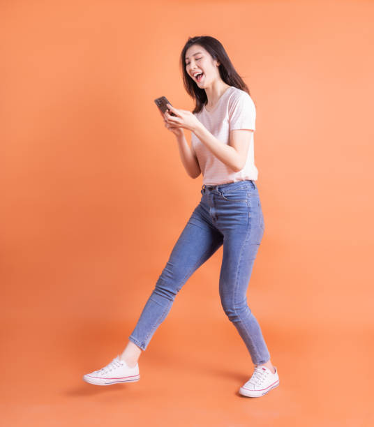 imagen de cuerpo entero de una joven asiática usando un teléfono inteligente sobre fondo naranja - female young women red hair full length fotografías e imágenes de stock