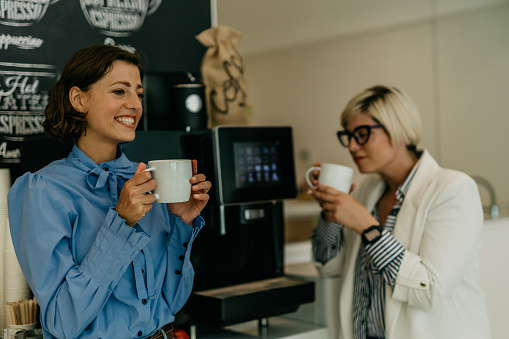 Image of two female coworkers in office holding a coffee cups, laughing and taking a break in the office pantry