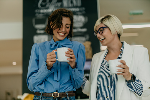 Spontaneous and loving photo of two beautiful young adults, business woman, enjoying their coffee brake in working space. Both smiling and chatting, a brunet and a blonde with eyeglasses on, hugging and having a moment of laughter while walking. Radiating positivity at work
