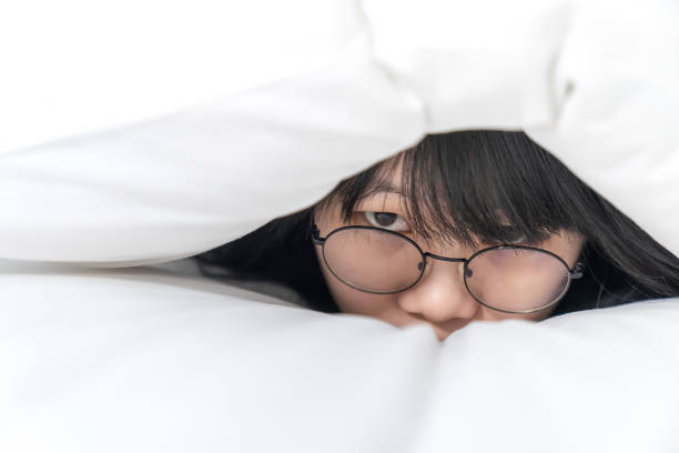 nahaufnahme der cute asian glasses frau, versteckt sie ihren körper in einem weißen kissen auf dem bett - hiding human eye fear women stock-fotos und bilder