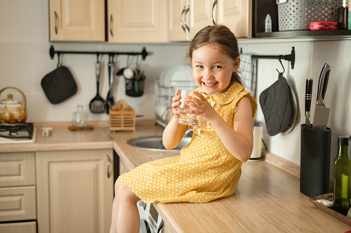 Beautiful smiling little caucasian girl drinking from glass of water against background of kitchen