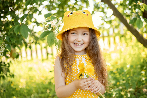 Summer portrait of cute beautiful smiling Caucasian girl in yellow hat and dress in park with bouquet of yellow dandelions in her hands