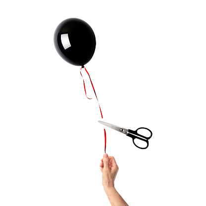 Scissors cutting the string of a hand-held a black color balloon against white background.