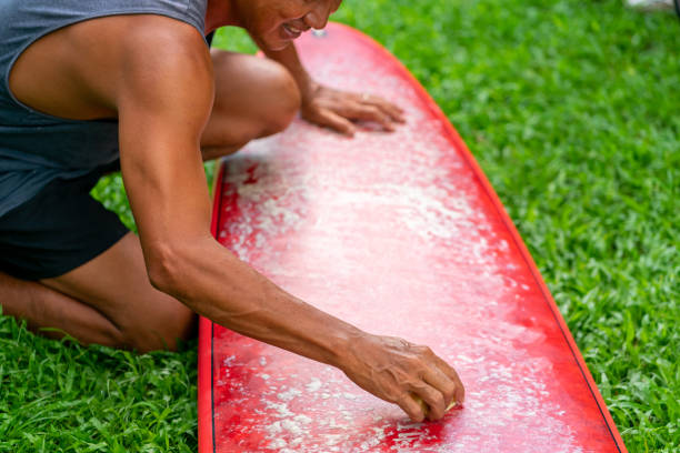 Healthy Asian senior man waxing surfboard surface at home. Active elderly male surfer enjoy outdoor lifestyle surfing at the sea on summer vacation. Retirement people activity and water sport concept.