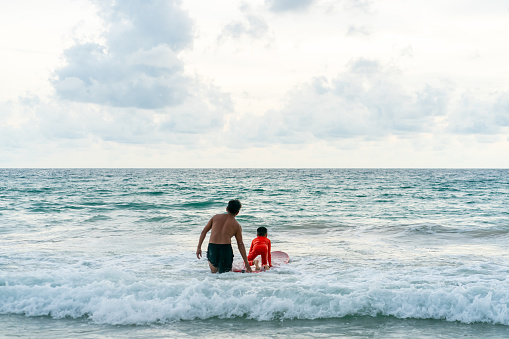 Happy Asian family grandfather teaching grandchild boy surfing on surfboard in the sea on summer vacation. Senior man and little boy enjoy outdoor activity lifestyle and water sport surfing together.