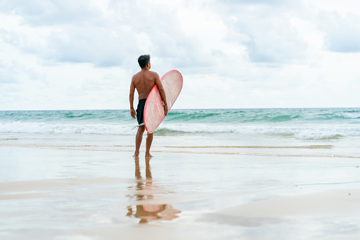Healthy Asian senior man surfer in swimwear holding surfboard walking on the beach at summer sunset. Elderly retired male enjoy outdoor activity lifestyle and water sport surfing on summer vacation