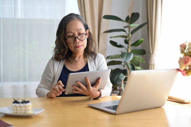 istock Concentrated middle aged woman sitting in front of laptop and using digital tablet while remote working from home. 1403405439