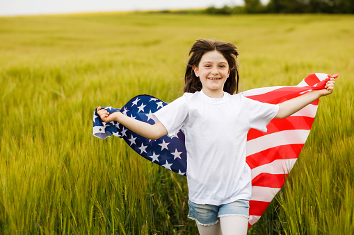 Carefree pretty girl running and smiling on the green field with a blowing flag of USA. Independence Day