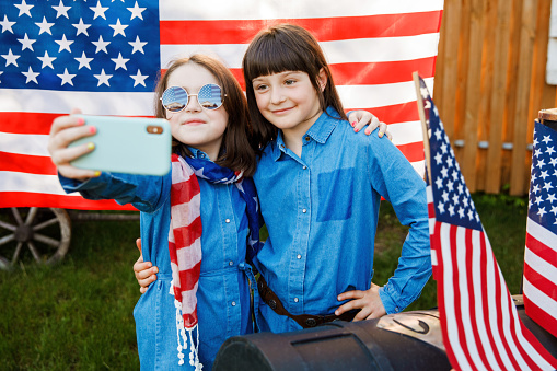 Two girls stand in the yard, holding the flag of USA and photographing themselves on a mobile phone. Independence Day
