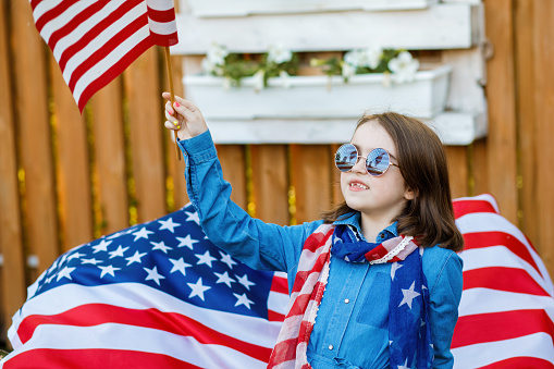 The girl stands in the yard, dressed in a denim dress and holding the flag of USA. Independence Day
