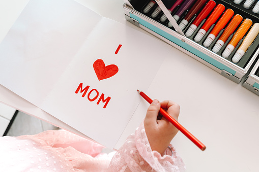 The child draws a card for Mother's Day. The girl writes on paper, I love you, mom.