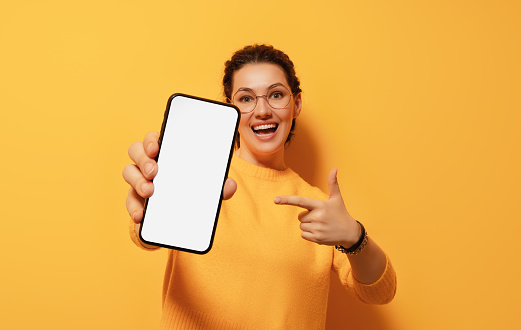 Happy young woman with smartphone on background of yellow wall. Blank screen mobile phone for graphic montage.