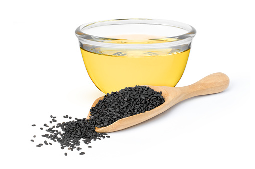 Sesame oil in glass bowl with black sesame seeds in wooden scoop isolated on white background.