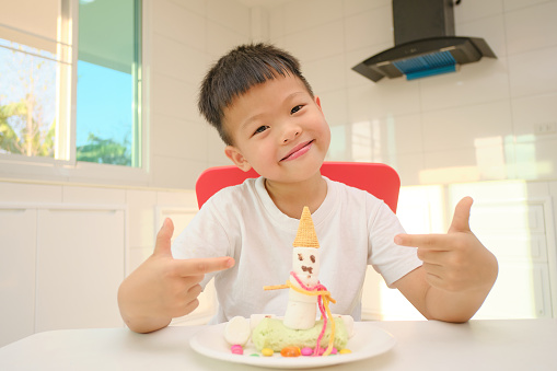 Cute little Asian kindergarten boy child sitting at the kitchen making easy and fun Marshmallow Snowman edible craft for kids, Montessori education, Homeschooling, Soft and selective focus
