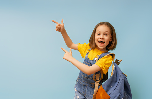 Cute happy kid standing on light blue background. Child with backpack, little girl is ready to back to school.