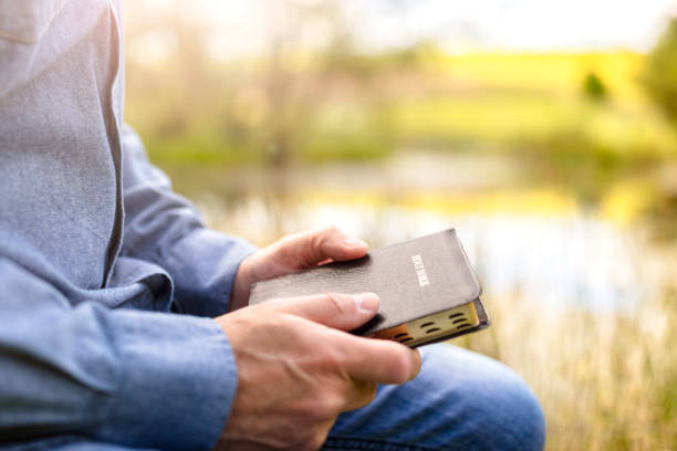 Christian man holding the Holy Bible at sunset by water and field background stock photo