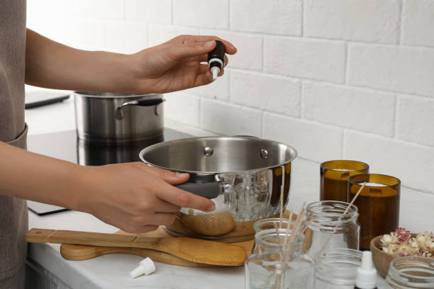 Woman adding essential oil into pot with melted wax at kitchen counter, closeup. Making homemade candles Woman adding essential oil into pot with melted wax at kitchen counter, closeup. Making homemade candles melting wax stock pictures, royalty-free photos & images