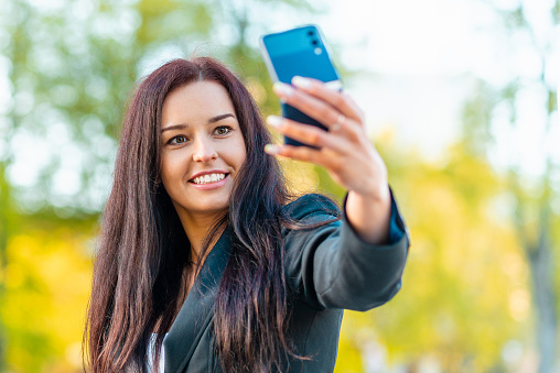 Portrait of a young beautiful business woman in a suit.Busineswoman walking through the city, video call, selfie, holding a phone.New generation business, communication.Blurred summer sunset.Close-up.