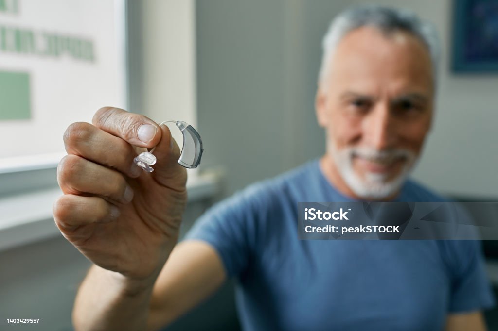 Senior man holding BTE hearing aid in hand on foreground, close-up. Treatment of deafness in elderly people Hearing Aid Stock Photo