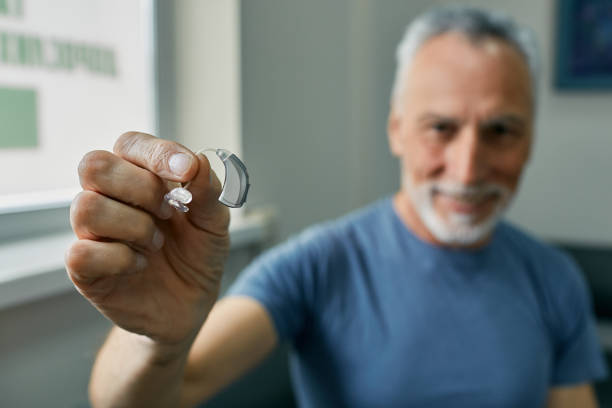 Senior man holding BTE hearing aid in hand on foreground, close-up. Treatment of deafness in elderly people stock photo