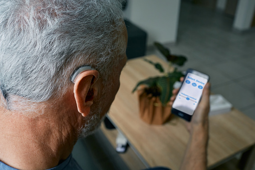 Hearing impaired senior man adjusting settings for his BTE hearing aid via smartphone. Hearing aids with innovative technologies at audiology