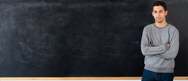 Thoughtful young arabic man standing next to empty chalk board at school or university, young male on blackboard background, knowledge concept, layout, banner