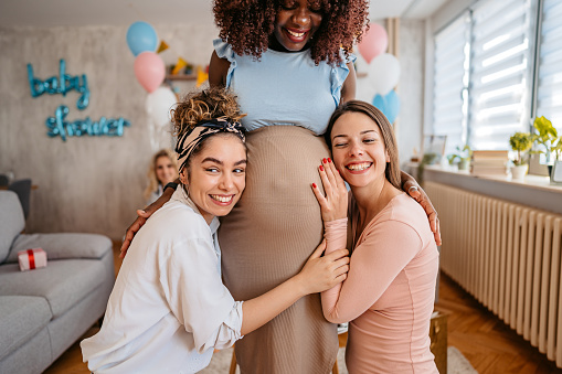 Beautiful young pregnant black woman having a baby shower with friends at home. Her two friends are touching her pregnant belly and embracing it.
