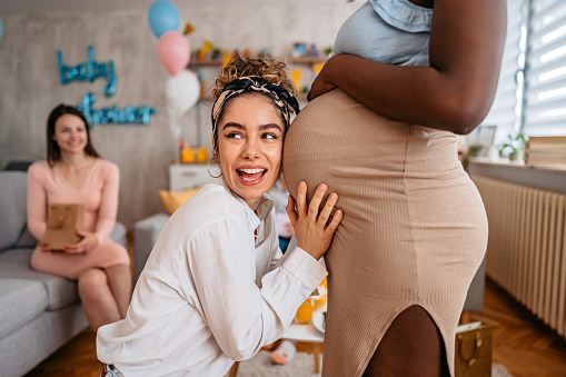 Beautiful young pregnant black woman having a baby shower with friends at home. Her friend is touching her pregnant belly and embracing it.