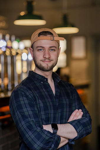 A portrait of a young man standing in a bar establishment in Newcastle upon Tyne, England. He is looking at the camera and smiling with his arms crossed.