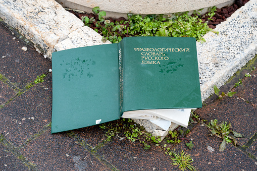 A thick book in hardcover lies on the street, thrown away on the cover is written in Russian - Phraseological Dictionary of the Russian Language. Close-up.