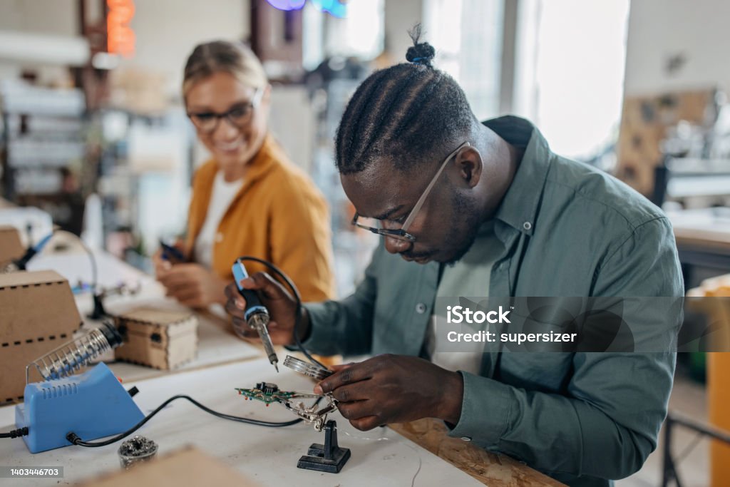 One more circuit board Ona male african American and one Caucasian female student working in the university workshop. He is soldering circuit board Computer Chip Stock Photo
