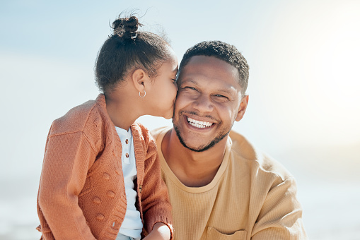 Adorable chica de raza mixta besa a su padre en la mejilla mientras está en la playa. Hija pequeña mostrando amor y afecto a papá mientras sonríe y mira a la cámara photo