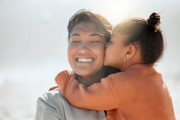 une adorable fille métisse embrasse sa mère sur la joue et tend ses bras autour d’elle à la plage. petite fille montrant de l’amour et de l’affection à maman pendant qu’elle ferme les yeux et sourit - boucle doreille photos et images de collection