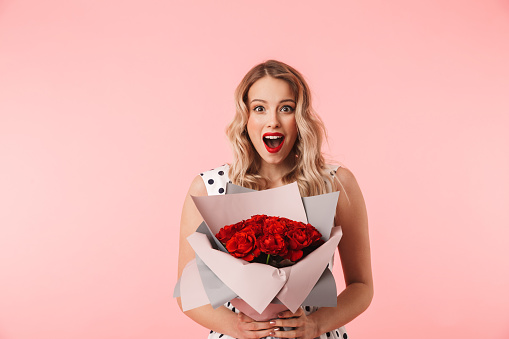 Beautiful young blonde woman wearing dress standing isolated over pink background, holding bouquet of roses