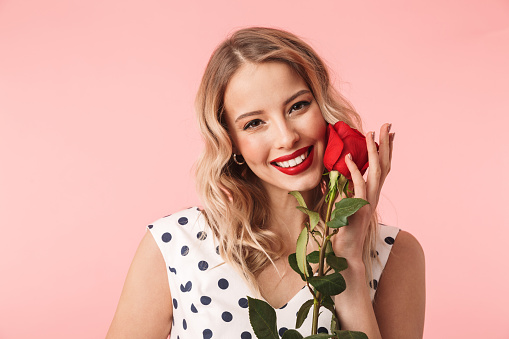 Beautiful woman with long blonde hair and red lips wearing pink clothes poses near blooming roses in garden. Wear light pink top and skirt