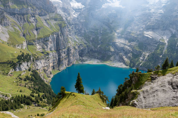 Panoramic view of the lake Oeschinensee View from the panoramic hiking trail of the mountain lake and its surrounding steep lake shores, in the foreground a mountain ridge with a single tree on it. The famous Oeschinensee lake which is located at 1500 meters above sea level and is located above Kandersteg in the Bernese Oberland, Canton of Bern, Switzerland lake oeschinensee stock pictures, royalty-free photos & images