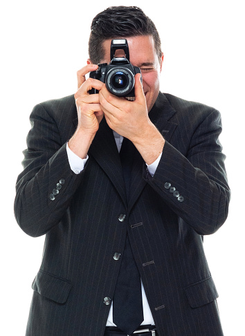 Front view of caucasian young male tourist standing in front of white background wearing businesswear who is laughing who is photographing and holding camera