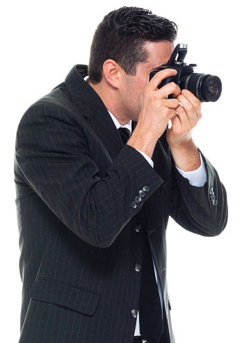 Profile view of caucasian male photographer standing in front of white background wearing businesswear who is laughing who is photographing and holding camera