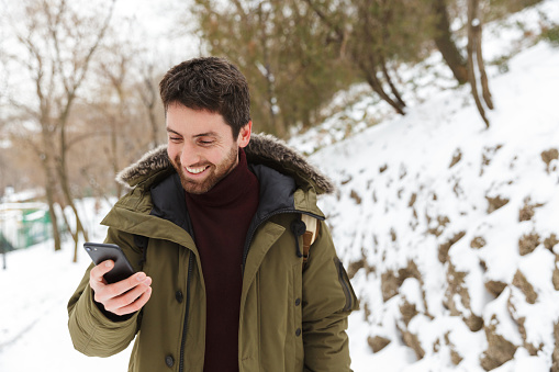 Handsome young man wearing winter jacket using mobile phone while walking outdoors