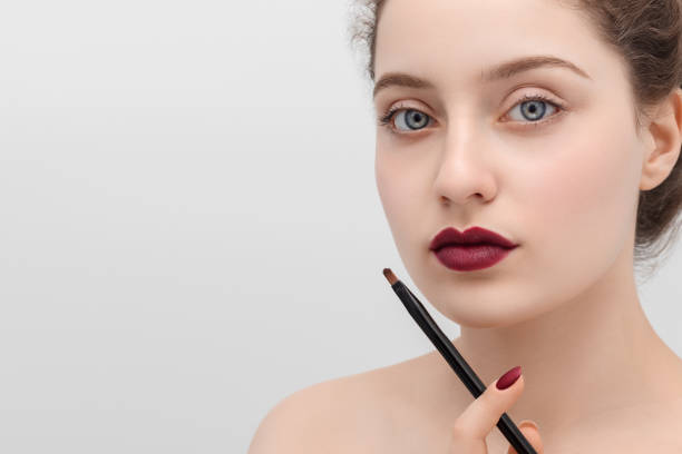 Close up shot of a beautiful young woman with blue eyes and dark red lipstick holding make up brush. Copy space on the left. stock photo