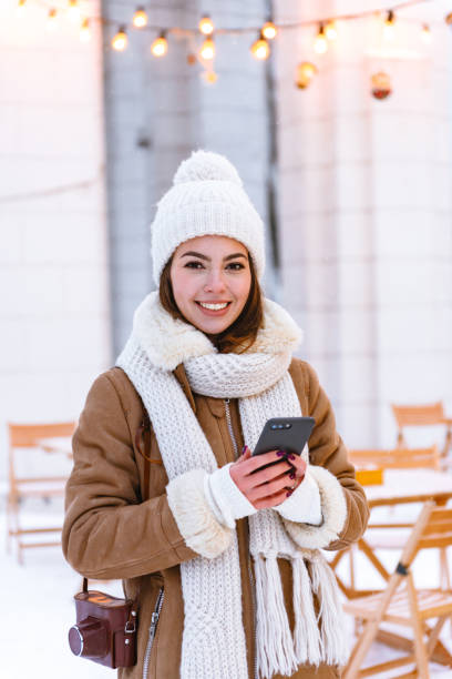 istock Young woman in hat and scarf walking outdoors in winter snow drinking coffee using mobile phone. 1403461506