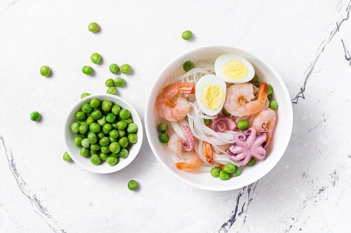 Top view of bowl with chinese rice noodles, tasty shrimps, bebe octopus, chicken eggs and green peas on white marble background