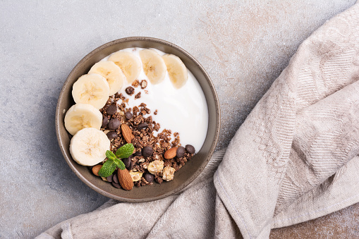 Top view of granola with yogurt, fresh banana, chocolate drops and almond nuts for healthy breakfast on grey concrete background