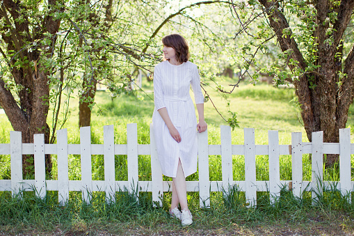 Beautiful caucasian woman in white dress walks in spring blossom garden near white wooden fence