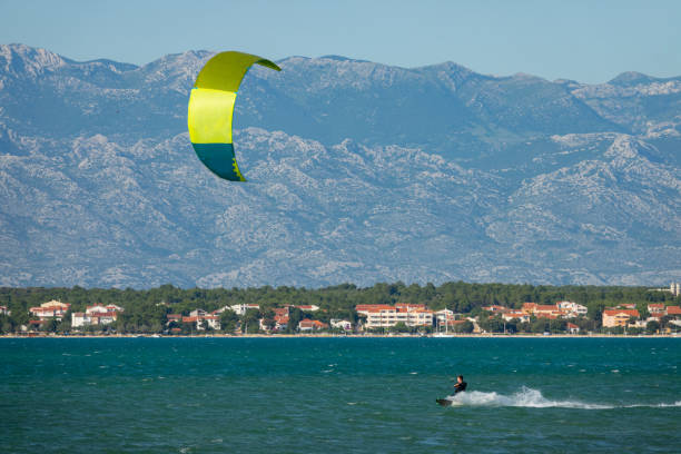 Man on active vacation in Croatia kitesurfs past a coast under the Velebit mountain range. Scenic action shot of a kitesurfer riding waves on a sunny day in the picturesque green Croatian seaside.