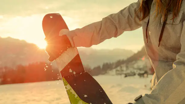 CLOSE UP, LENS FLARE, DOF: Active tourist rips skins off the bottom of their split boarding gear before riding at sunrise. Split boarder takes the protective layer off the bottom of their new skis.