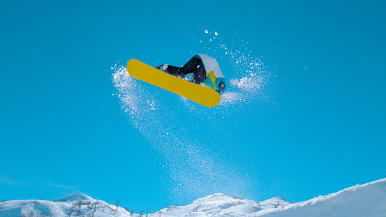 BOTTOM UP: Athletic male tourist snowboarding in the sunny Julian Alps does a spinning trick as he takes off a massive kicker. Bottom up action shot of a pro doing spectacular snowboarding stunts.