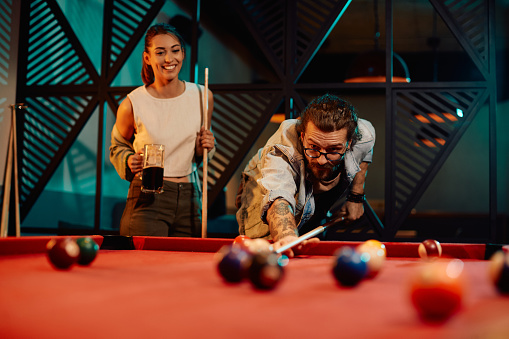 Young man playing snooker with his girlfriend and aiming at the ball.