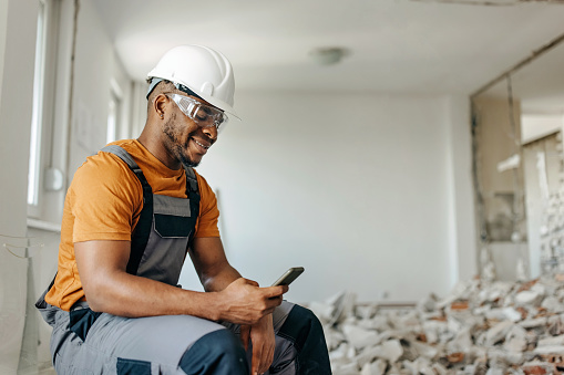 An African American man taking a social media break from his home renovation project, sitting on a chair, smiling at his cellphone.