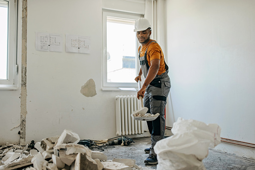 An African American male construction worker in a protective uniform standing in a room, putting away residue with a shovel.